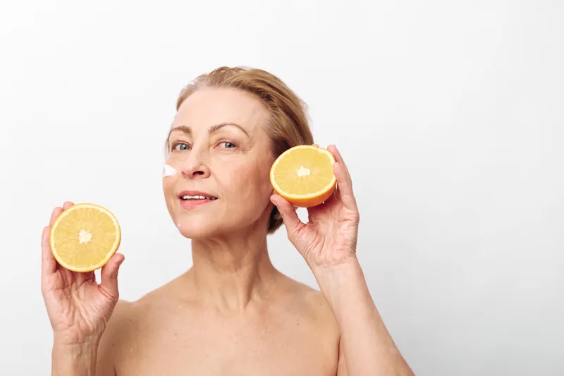 Mature woman with bare shoulders holds two halves of fresh grapefruit near her face. Bright white background highlights healthy skin and natural fruit for wellness and skincare concept.
