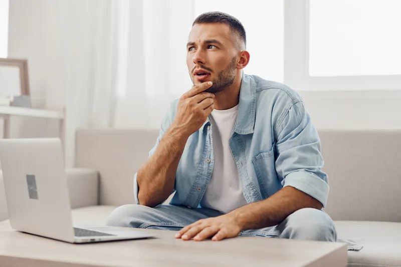 Thoughtful man pondering ideas at home with a laptop, showcasing a relaxed yet focused atmosphere in a modern living space, expressing curiosity and creativity.