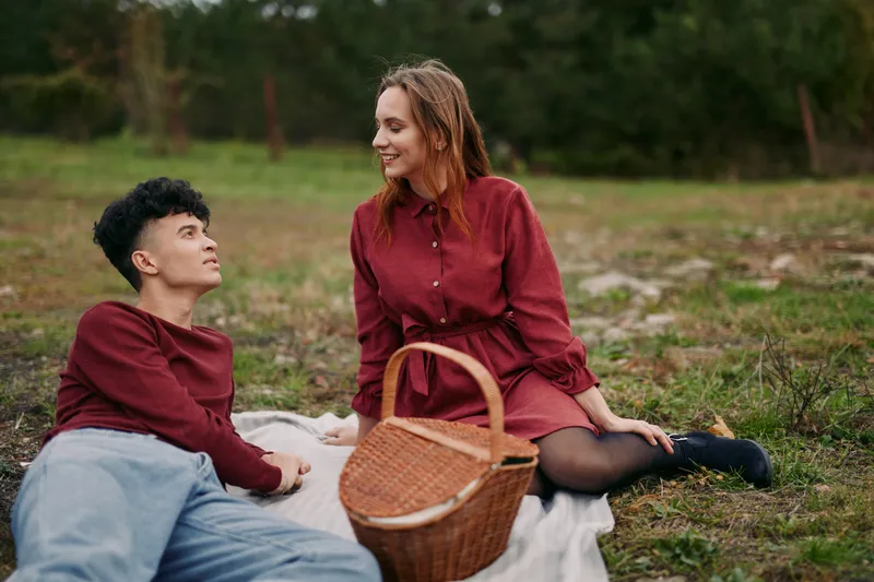 A candid outdoor picnic scene capturing an authentic moment between two young adults sharing a genuine connection on a blanket, natural expressions and realistic interaction in soft daylight.