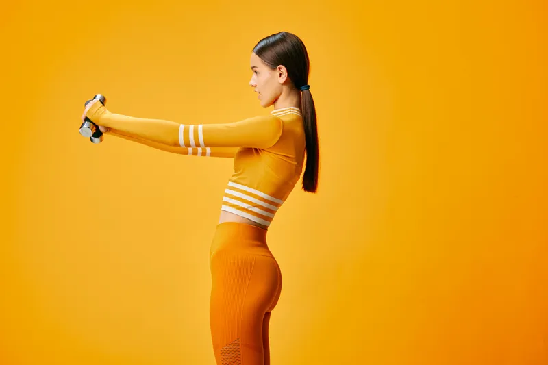 Woman fitness portrait: sporty female in orange workout set holds dumbbells in front, side profile with ponytail, focused expression and strong posture against vivid orange studio background.