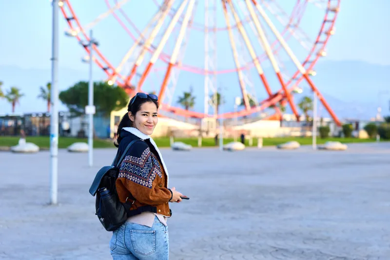 Woman with backpack in outdoor amusement park near a towering ferris wheel, casual fashion style, evening light, cheerful mood, urban travel vibe and wide blue sky
