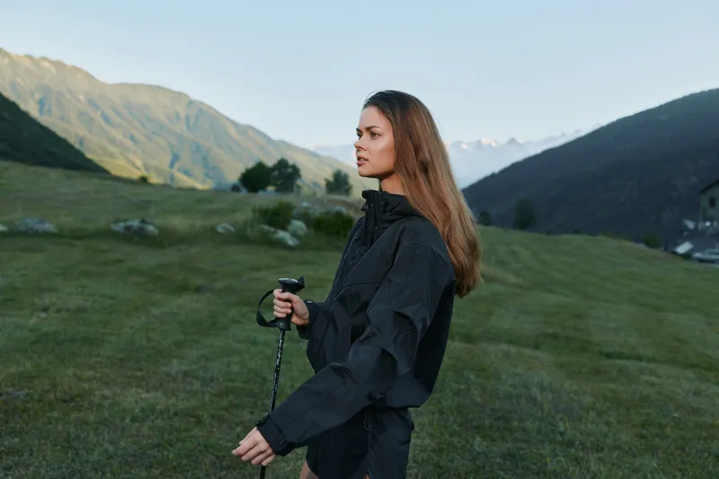 A woman stands in a grassy valley with distant mountains, holding a camera and monopod, dressed in a windproof jacket, ready for outdoor photography in a calm dawn light.