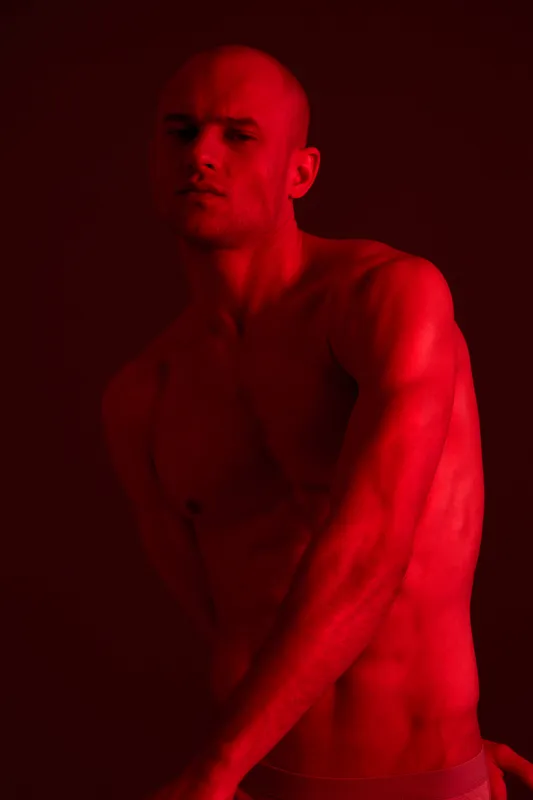 Strong man posing under dramatic red light, showcasing muscular physique and intense expression. This black and white photograph highlights strength and power.