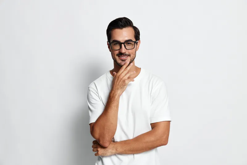 Handsome man in casual white t-shirt and glasses smiling confidently while looking at the camera with thoughtful expression isolated on plain light background.