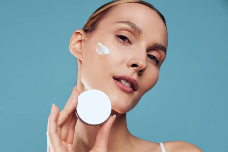 Young woman applies face cream on cheek holds cosmetic jar close to skin demonstrates skincare routine with natural makeup and smooth healthy complexion on blue background.