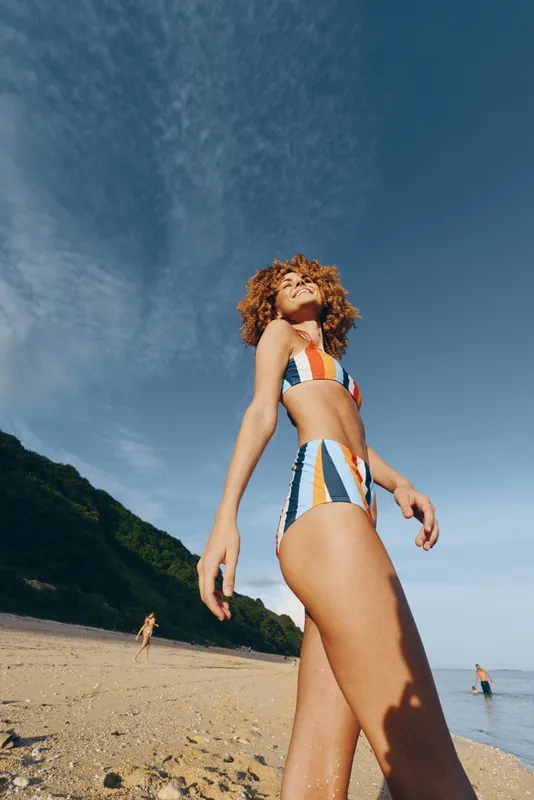 Curly haired woman in striped swimsuit walks along sandy beach toward ocean under bright summer sunlight distant green hills behind casual vacation mood natural skin tones outdoors travel