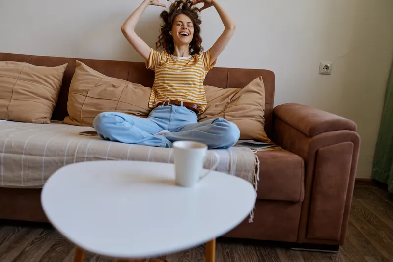 Young woman relaxes at home on a sofa, casual striped shirt and jeans, cheerful mood, coffee cup on a small table, bright living room backdrop, lifestyle portrait