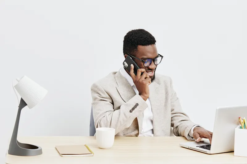 Professional young African American man wearing glasses, beige blazer, working at desk while talking on phone and using laptop, minimalistic office background, business and communication concept