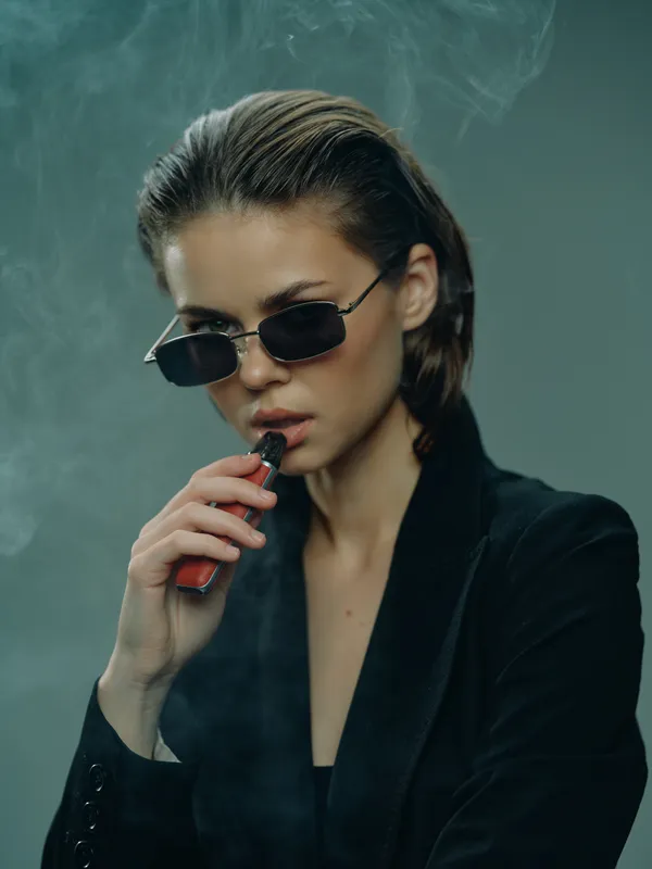 Stylish woman vaping in black suit and sunglasses with smoky background, expressing cool and modern attitude in studio setting.