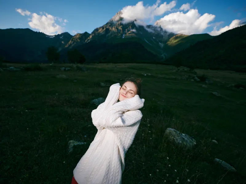 Woman in a cozy white sweater hugs herself in a wide meadow with mountains and a bright sky, conveying outdoor happiness, serenity, and personal wellbeing