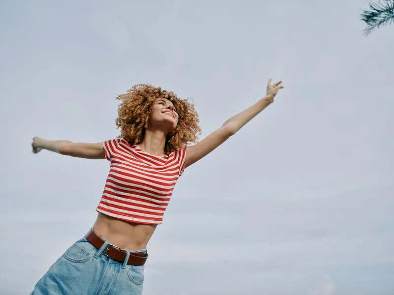 Joyful woman outdoors with arms wide, wearing striped crop top and denim jeans, basking in sunlight, embodying freedom, balance, and carefree energy against a clear blue sky.