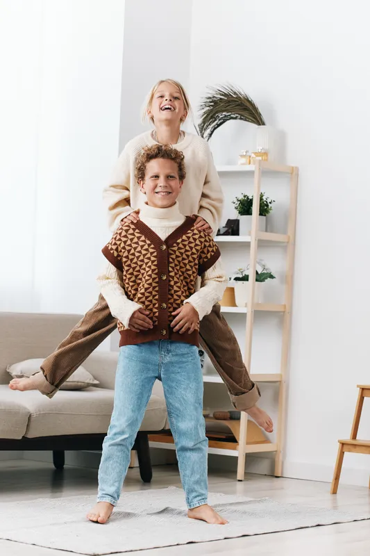 Two joyful children share a playful piggyback moment indoors, a bright living room scene capturing carefree laughter, connection, and a candid family playful dynamic.