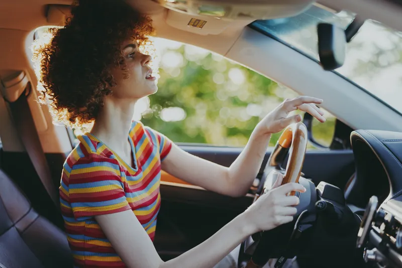 A woman driver with curly hair sits behind the wheel in a striped tee, daylight streams through the car, capturing casual road trip mood and retro lifestyle.
