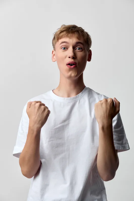 Excited, surprised young man raises fists in a white tee, capturing energetic emotion and a triumphant mood, ideal for lifestyle, youth and celebration themed campaigns