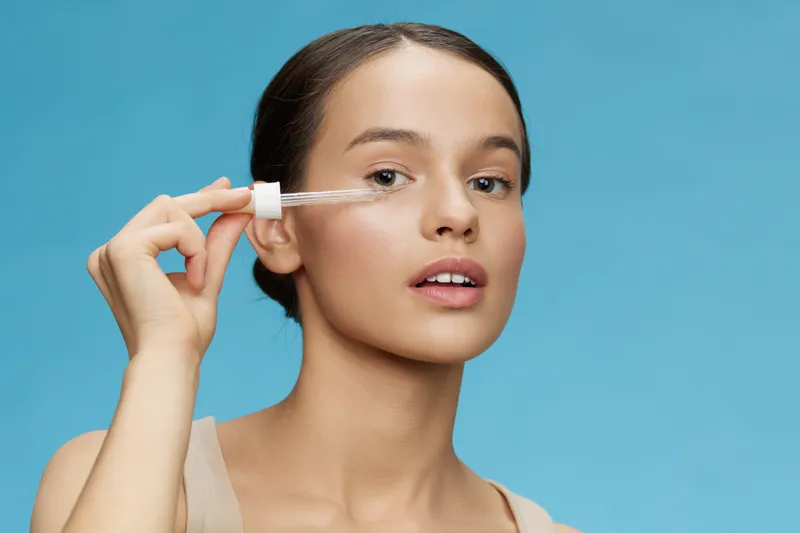 Young woman applying serum on her face with a dropper, highlighting skincare routine, healthy skin, and beauty concept against a blue background