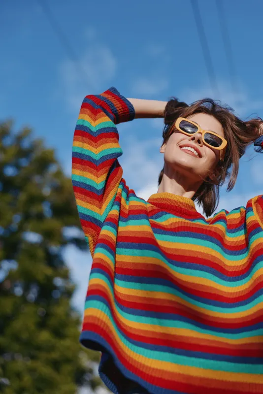 Woman rainbow sweater sunglasses smiling outdoors portrait of joyful young person under blue sky, colorful knit fashion, casual jumper and trendy eyewear conveying carefree summer mood.