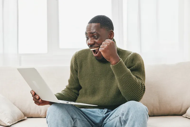 Excited young man celebrating success while using a laptop at home, showcasing emotions of joy and achievement in a bright, cozy setting