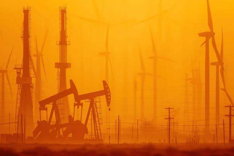 Драматичный закат окрашивает нефтяные качалки и ветряные турбины в теплый оранжевый свет, символизируя контраст между традиционными и возобновляемыми источниками энергии