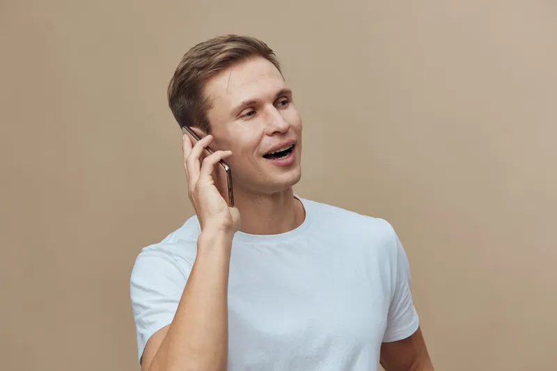 Happy young man with short brown hair in casual white t-shirt talking on mobile phone smiling isolated on plain beige background studio portrait. People lifestyle concept with cheerful mood