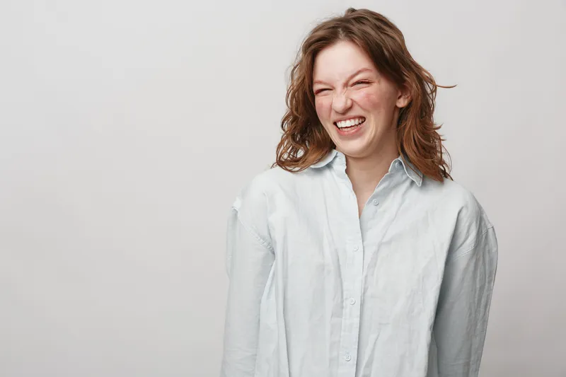 Happy young woman with curly hair laughs joyfully wearing white shirt on light background. Concept of positive emotions, happiness, and natural beauty in casual style.