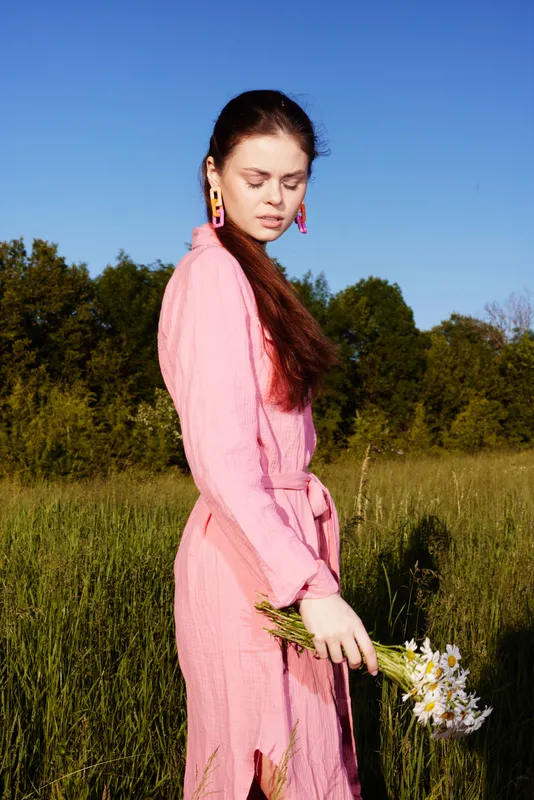 A young woman in a pink dress gracefully holds flowers in a vibrant green meadow under a clear blue sky, embodying spring s elegance and joy