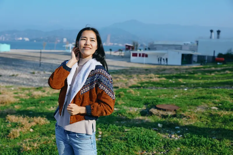 Smiling woman in a knitted jacket and scarf stands on a grassy coastal field with sea and hills in the distance, conveying casual style, outdoor lifestyle, and carefree moment.