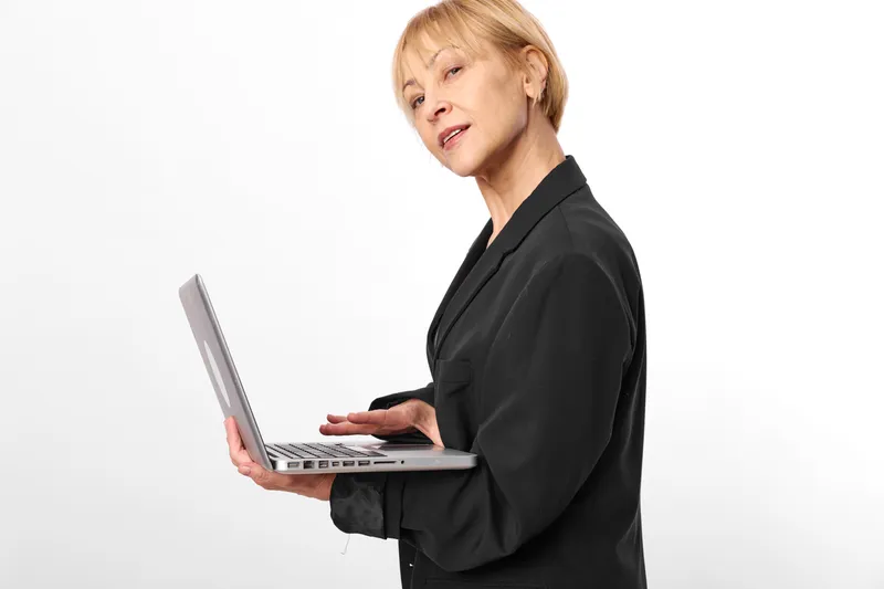 Mature woman in black suit holding a laptop, speaking with concern in a studio, remote work concept, business communication, modern technology, isolated white background, professional