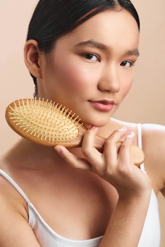 Asian woman holds a wooden hair brush near her face, looking softly at the camera with a calm expression, in a neutral studio setting highlighting grooming and personal care.