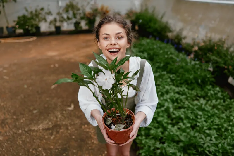Joyful young woman with a potted plant in a greenhouse, celebrating nature and gardening, wearing casual clothes in a vibrant green setting
