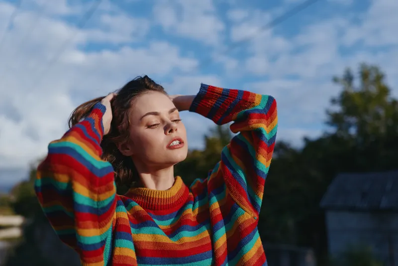 Woman portrait in a rainbow sweater outdoors, relaxed with eyes closed and sunlight on her face, hands in hair, colorful fashion and cozy seasonal style in natural countryside setting.