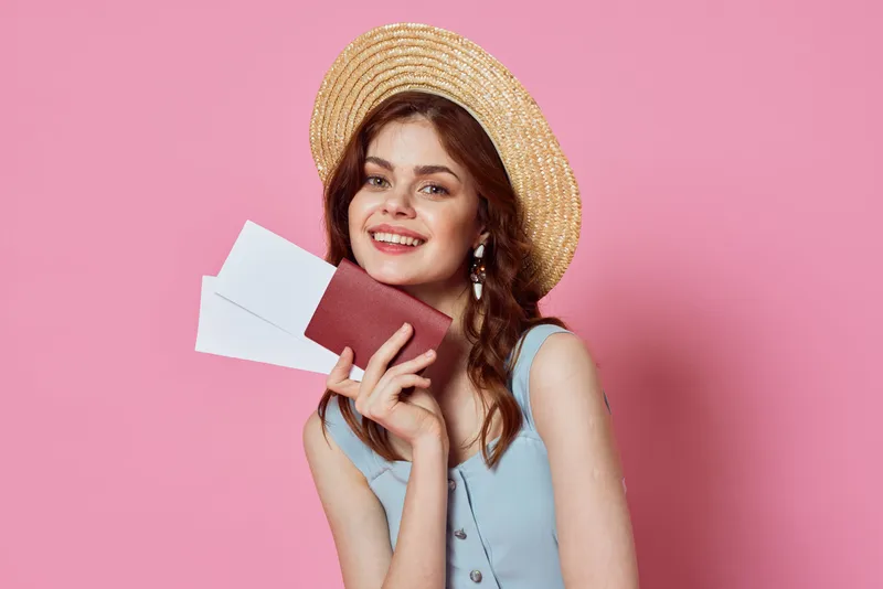Женщина в шляпе держит паспорт и авиабилет на фоне розового цвета, создавая образ пассажирки аэропорта