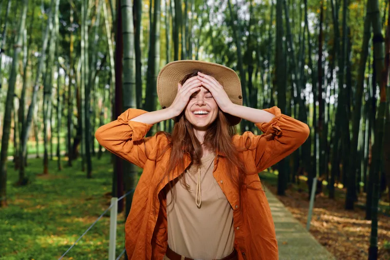Woman hat bamboo forest smiling walkway nature portrait of a joyful traveler wearing casual jacket on a wooden path, hands on face expressing relaxed happiness in green outdoor scenery.