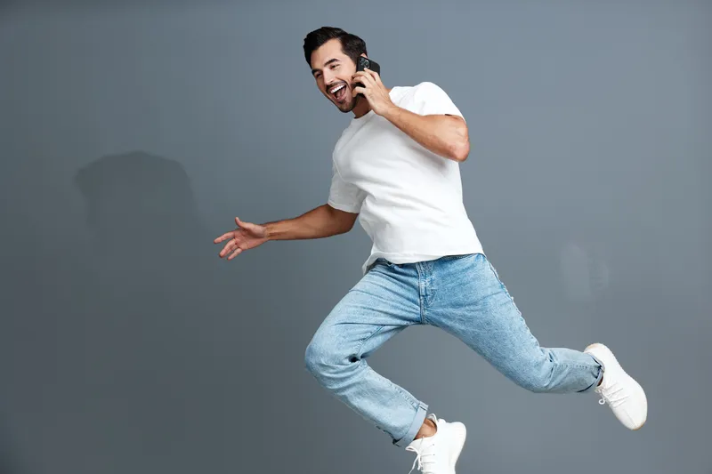 Full body joyful happy young man wearing white t-shirt jumping while talking on mobile phone against a bright gray backdrop. Lifestyle concept.