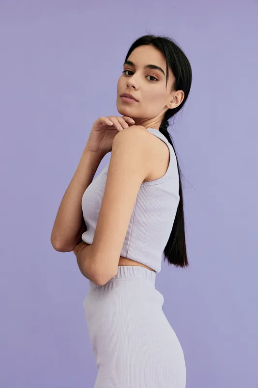Young woman with long dark hair wearing a light purple knitted sleeveless top and skirt, posing sideways against a pastel lavender background in soft natural lighting.
