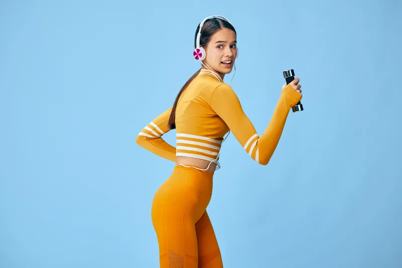 Fitness woman in vibrant orange athletic wear poses mid workout with dumbbells against a blue background, showcasing strength, balance and energetic movement for sport and wellness images.