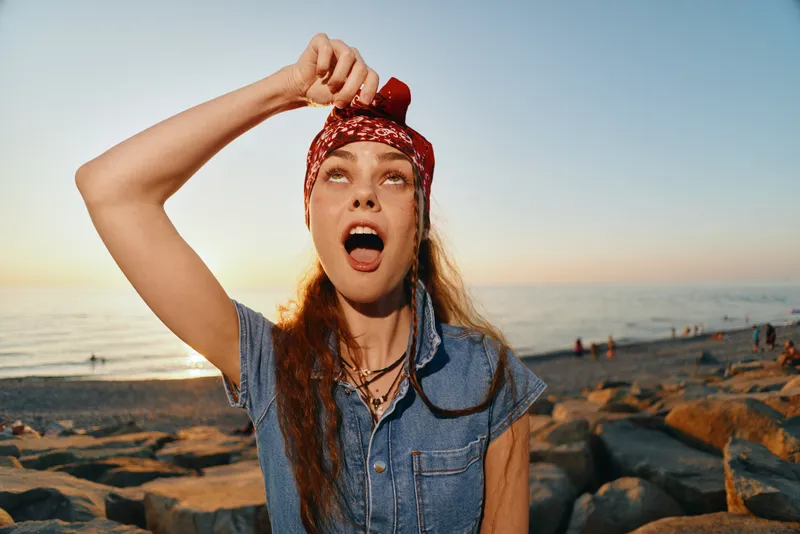 lifestyle woman in modern boho-western denim street style posing outdoors with film color effect under warm sunset light by the rocky beach