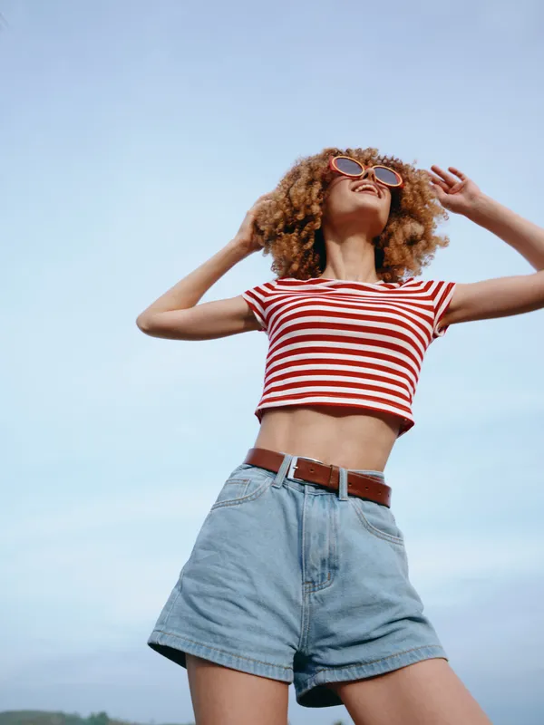 Vibrant portrait of a young woman in a red and white striped crop top and denim shorts, dancing outdoors under a clear blue sky with carefree energy and movement.
