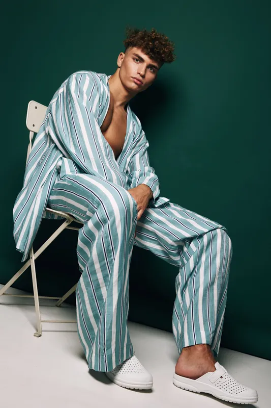 Young man sitting on chair wearing striped loose shirt and pants with white slip-on shoes against dark green background in studio. Fashion, casual style and relaxed pose.