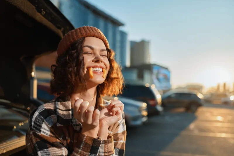 Woman smile vintage cars golden hour candid beanie: joyful young woman in a beanie and plaid coat laughing with candy in her mouth beside vintage cars at sunset, authenticity and golden hour glow.