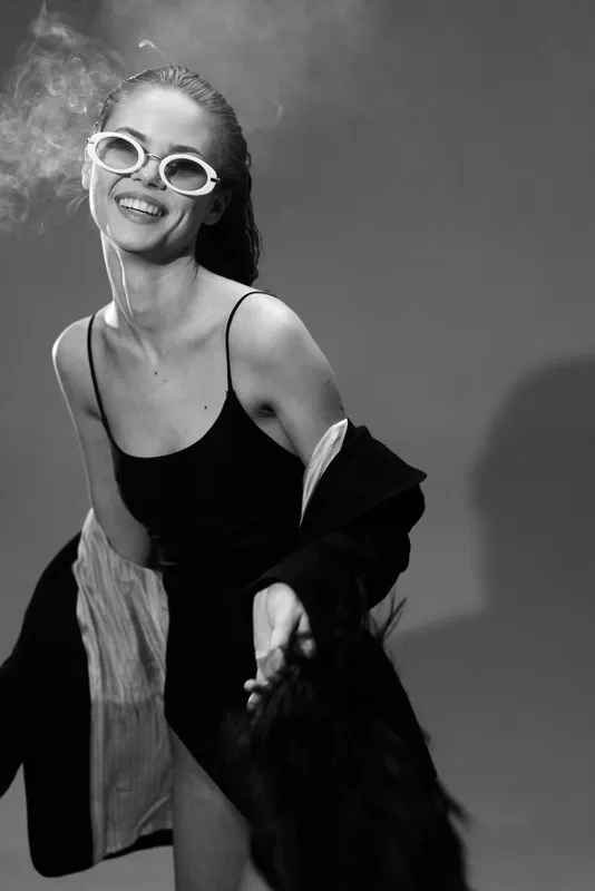 Smiling woman wearing sunglasses in a black dress with jacket, joyful and stylish, black and white portrait in studio lighting.