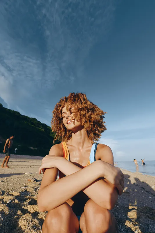 Cheerful outdoor portrait of a smiling woman with curly hair on a sunny beach, sunlit moment, relaxed posture, colorful swimsuit, clear blue sky, calm sea in the background