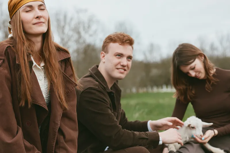 Three young adults enjoy a relaxed moment outdoors with a small baby goat, sitting on grass in casual autumn clothing and sharing a peaceful connection with nature.