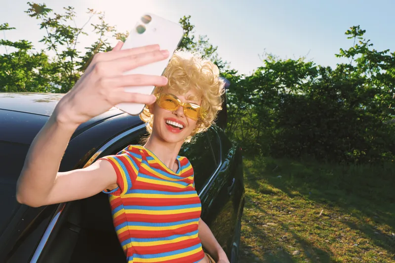 Cheerful woman outdoors, selfie by a car, striped shirt, sunlit moment, relaxed pose, retro film color vibe, vibrant greens, candid lifestyle scene, joyful energy