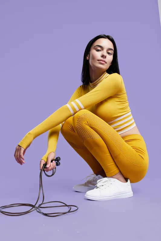 Sport fitness woman wearing yellow activewear squats holding jump rope on purple background, focused young female athlete training indoors with white sneakers.