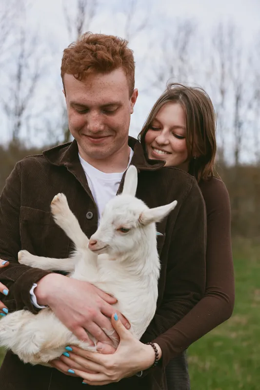 Young couple outdoors enjoying a tender moment while holding a white baby goat in a natural grassy field during a calm day in early spring.