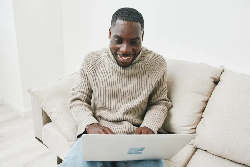 Happy Black man working on a laptop at home, wearing a cozy sweater, smiling as he enjoys his time and engages in remote work for a modern lifestyle