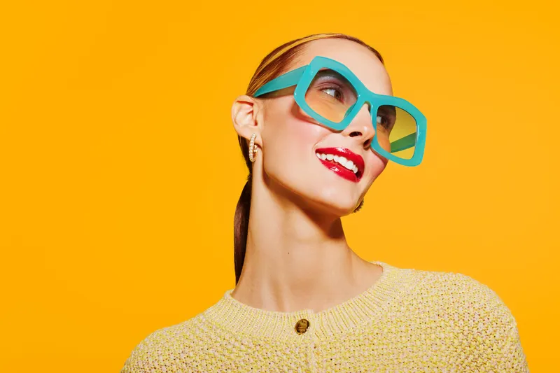 Stylish woman with bright red lipstick and turquoise sunglasses smiles confidently against vibrant yellow background. Concept of fashion, beauty, and summer style.