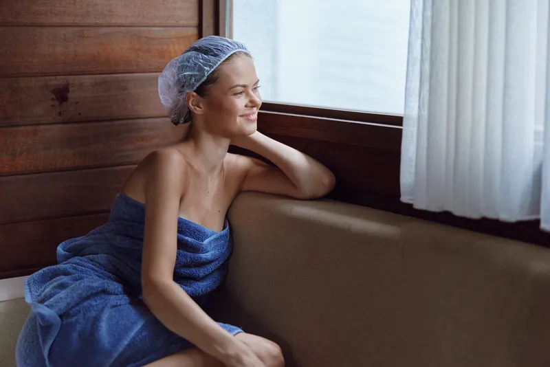 Smiling woman wrapped in blue towel resting by wooden wall and window, wearing shower cap, enjoying peaceful moment with natural light and cozy atmosphere indoors.
