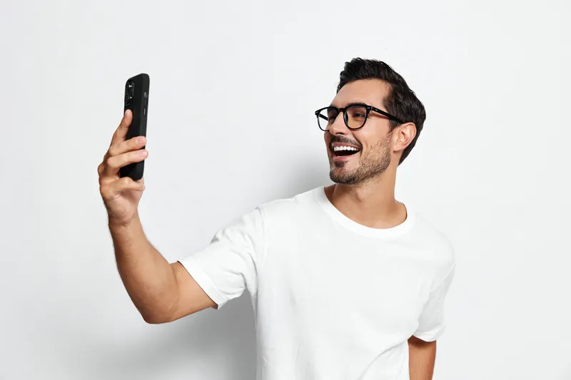 Smiling man wearing glasses holds smartphone taking selfie, cheerful expression on face against plain light background. Joyful moments concept.