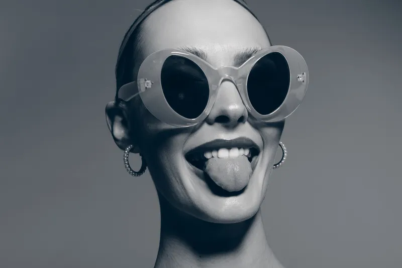 Closeup of white woman with pale skin wearing large round sunglasses and earrings, playfully sticking out tongue. Studio portrait with edgy and humorous concept of personality and style.